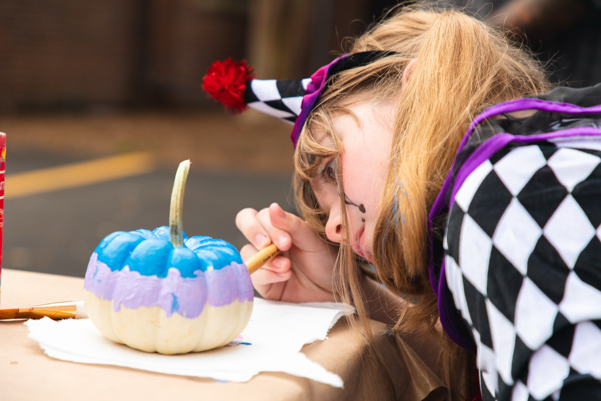 A kid in a harlequin costume carefully paints a small pumpkin