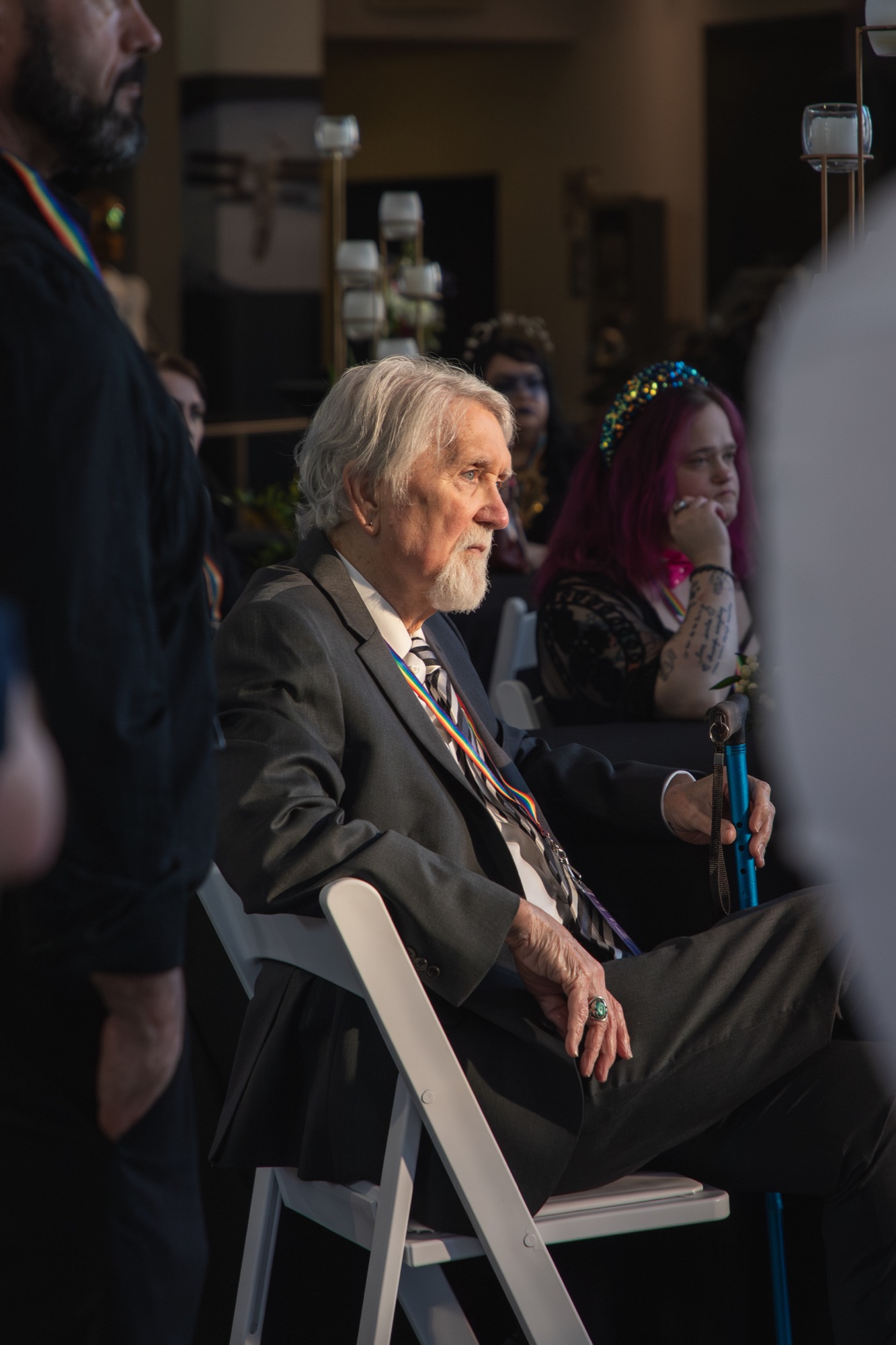 An older man in a suit and rainbow lanyard, lit by a shaft of window light